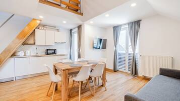 Une cuisine moderne avec des meubles blancs, une table à manger en bois avec quatre chaises, un téléviseur fixé au mur et un escalier avec une rampe en bois.
