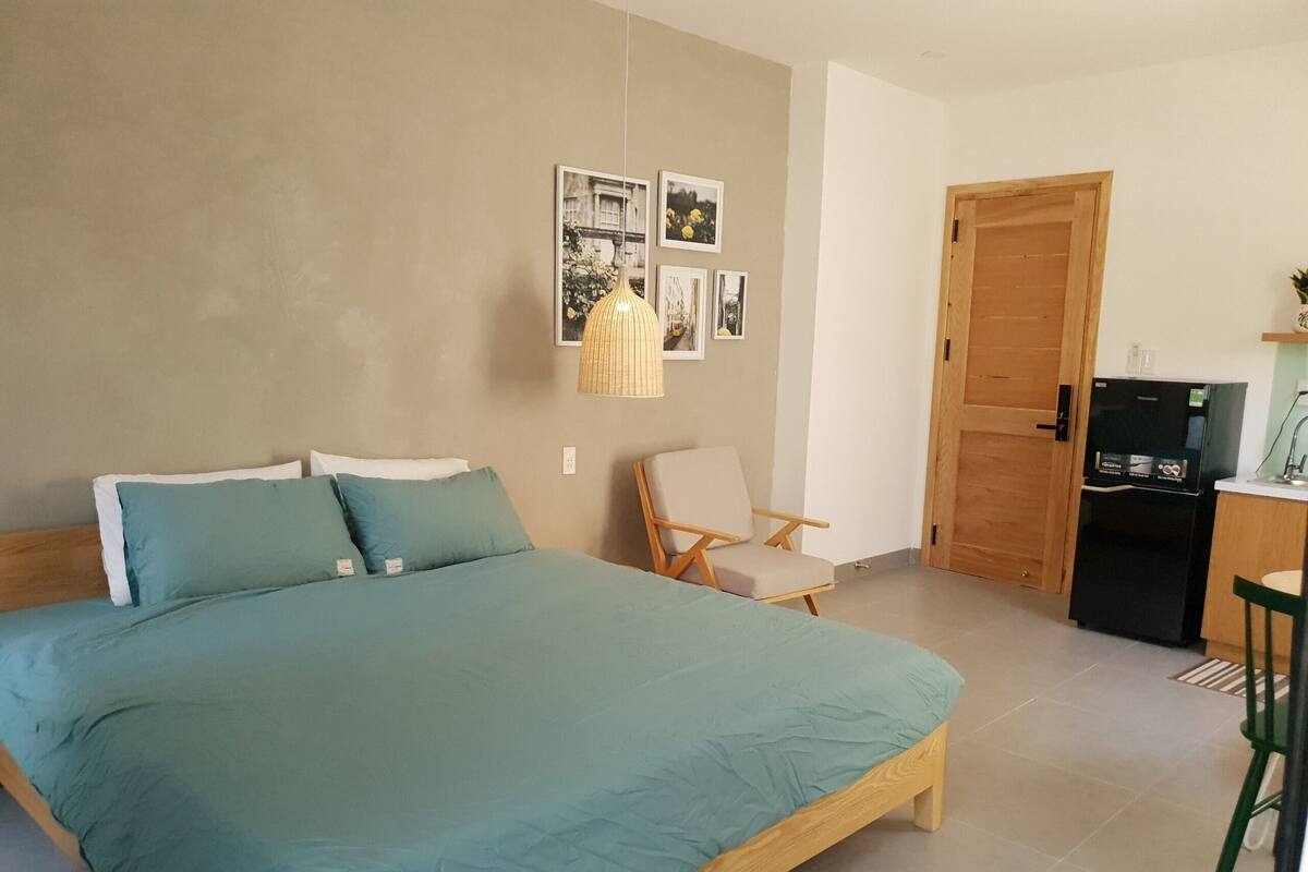 A bedroom with a bed, a chair, a desk with a refrigerator, and a door.