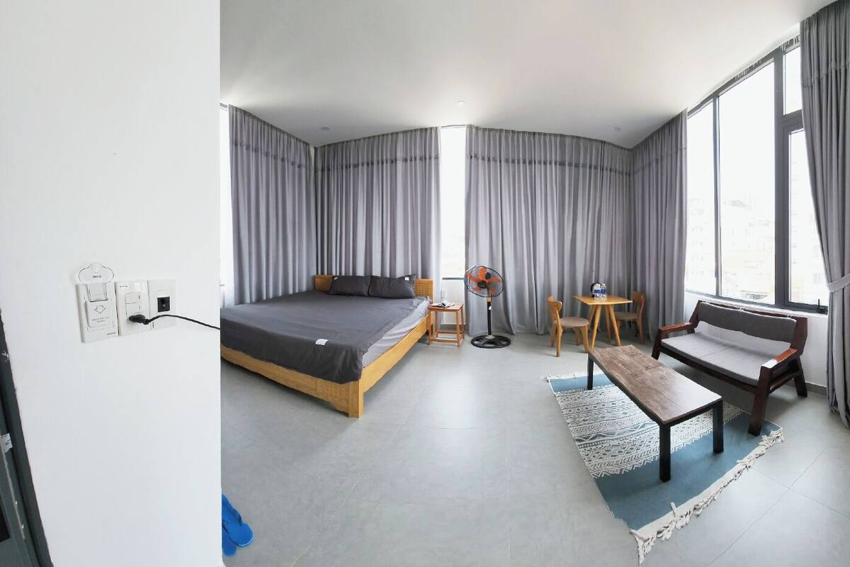 A modern hotel room with a bed, a small table, a bench, a fan, and a window with curtains.