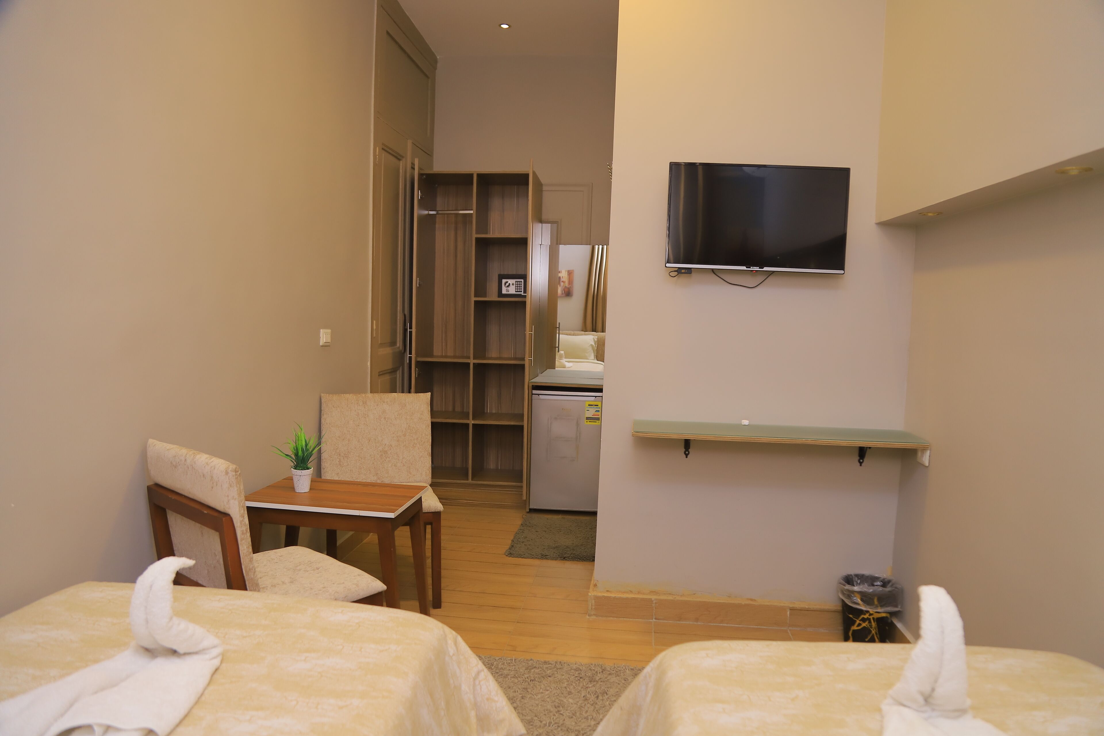 A hotel room with two beds, a TV mounted on the wall, a small table with a chair, a desk, and a shelf.