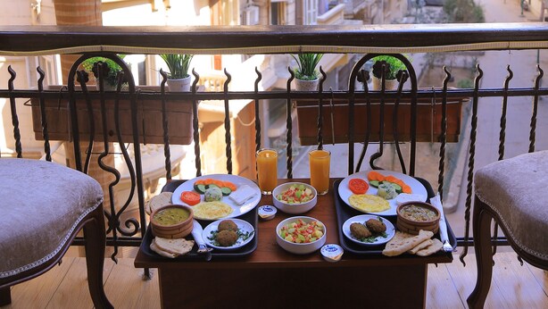 A balcony with a dining table set with food and drinks, overlooking a city street with buildings and palm trees. - Holy Sheet Hostel (Cairo)