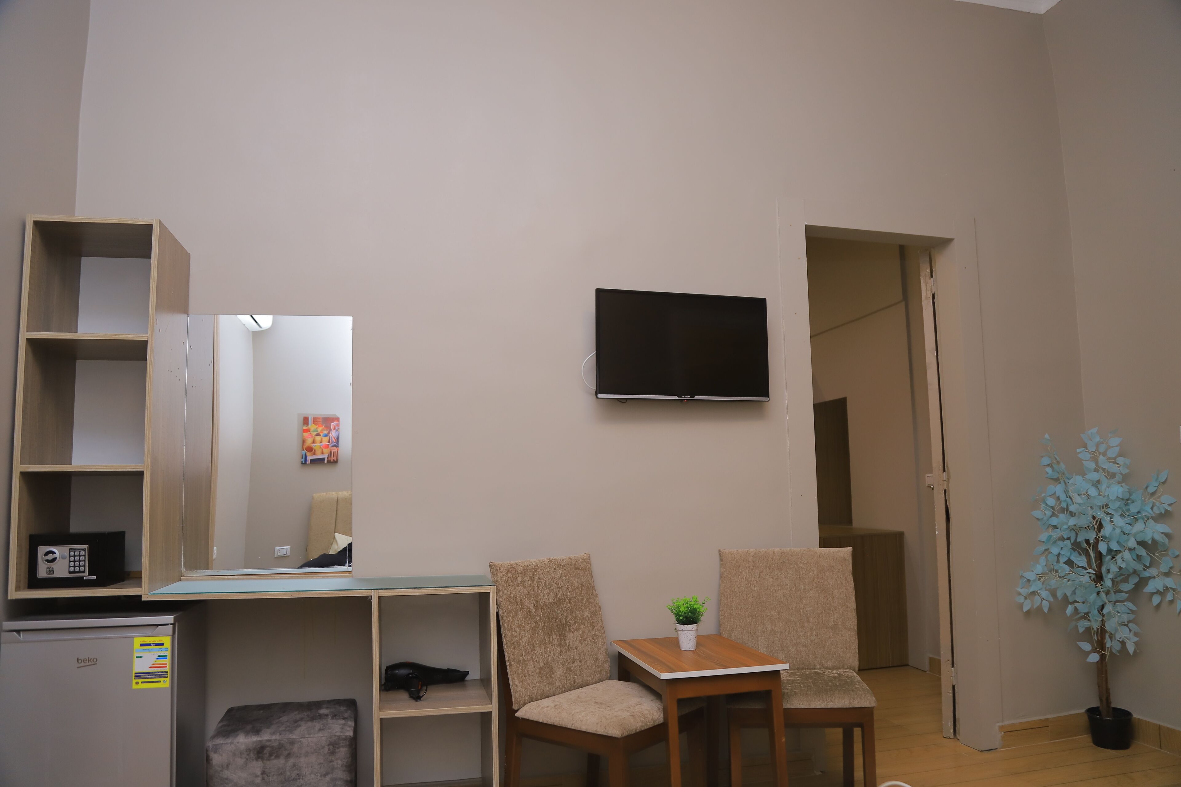 A room with a TV mounted on the wall, a small table with two chairs, a potted plant, and a refrigerator.