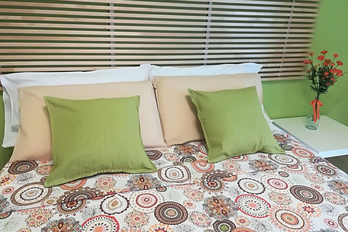 Una cama con una colcha estampada, dos almohadas verdes y un florero con flores sobre una mesita de noche.