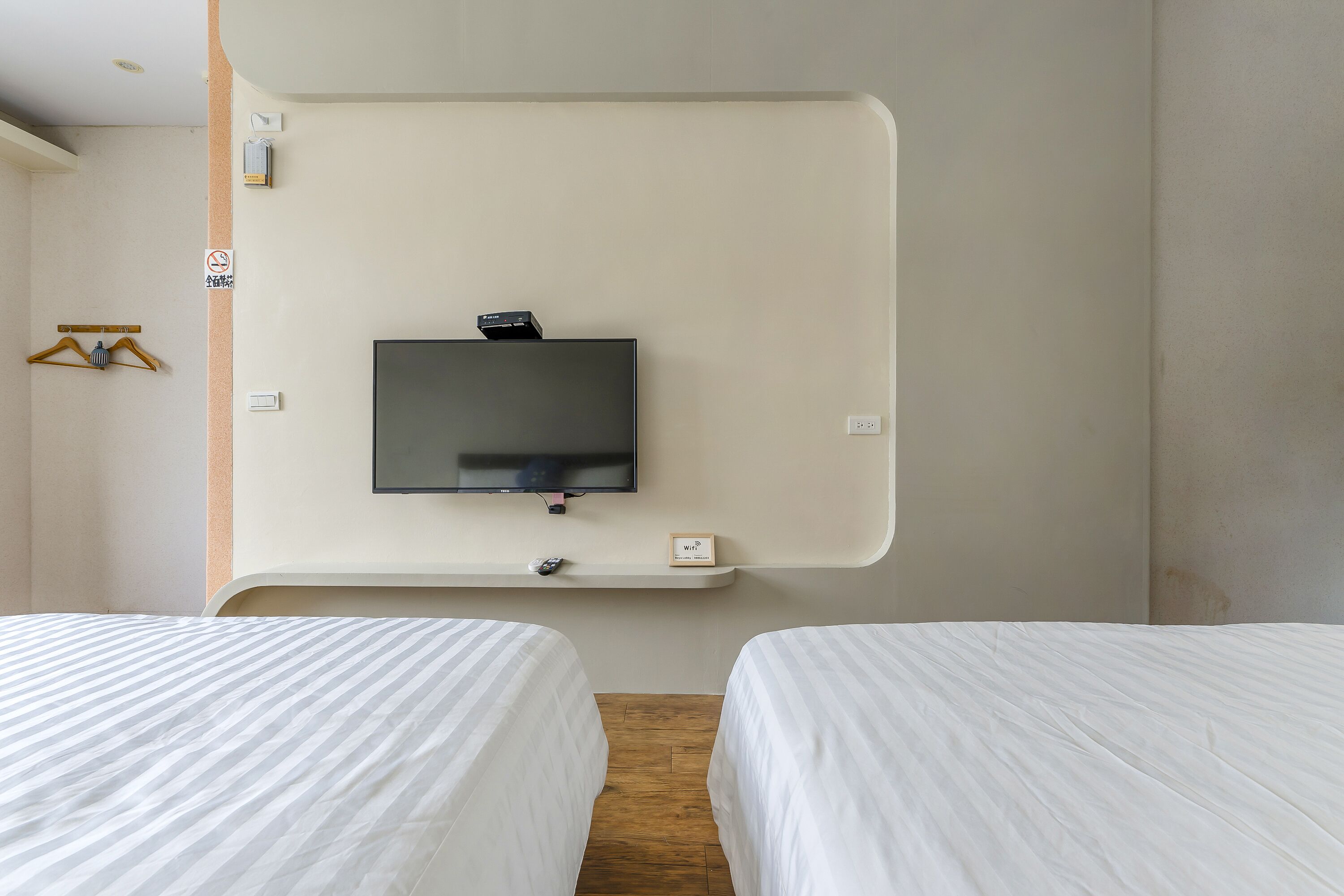 Habitación de hotel con dos camas, un televisor montado, un pequeño estante con una placa y suelo de madera.