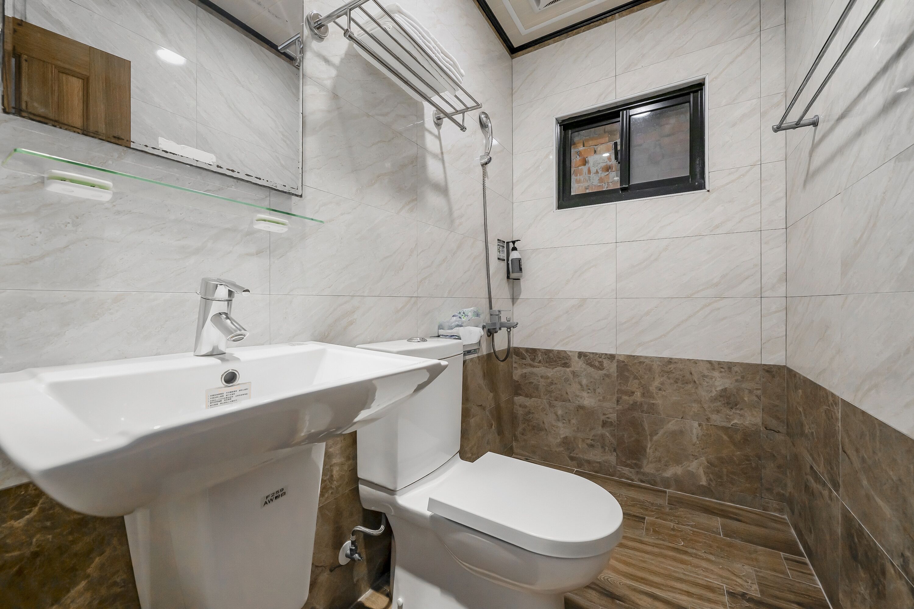 A bathroom with a white sink, toilet, and shower. There is a small window and a wooden cabinet above the sink.