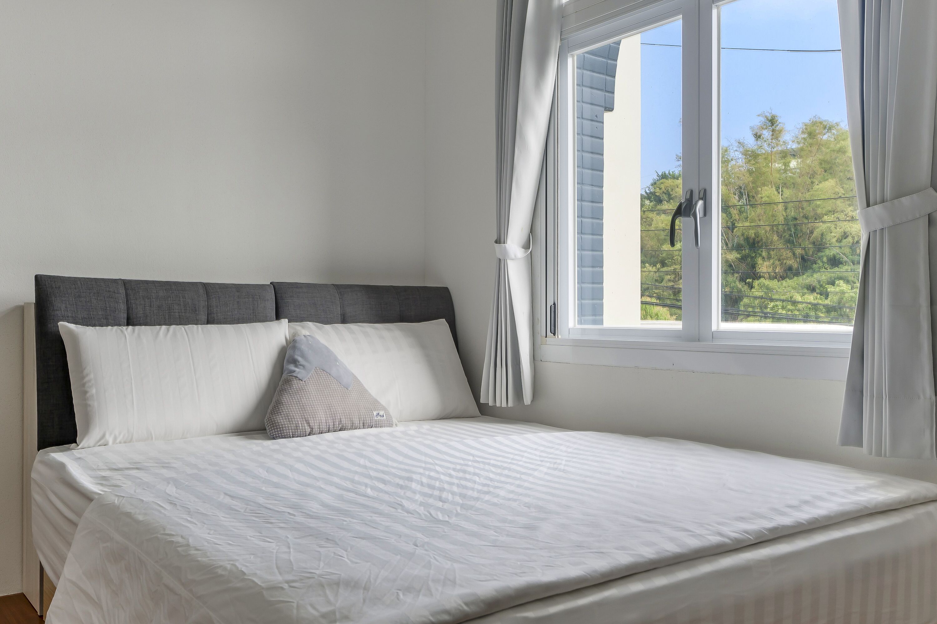 Una cama bien hecha con ropa blanca y un cabecero gris, una ventana con cortinas, y vista a zonas verdes al exterior.
