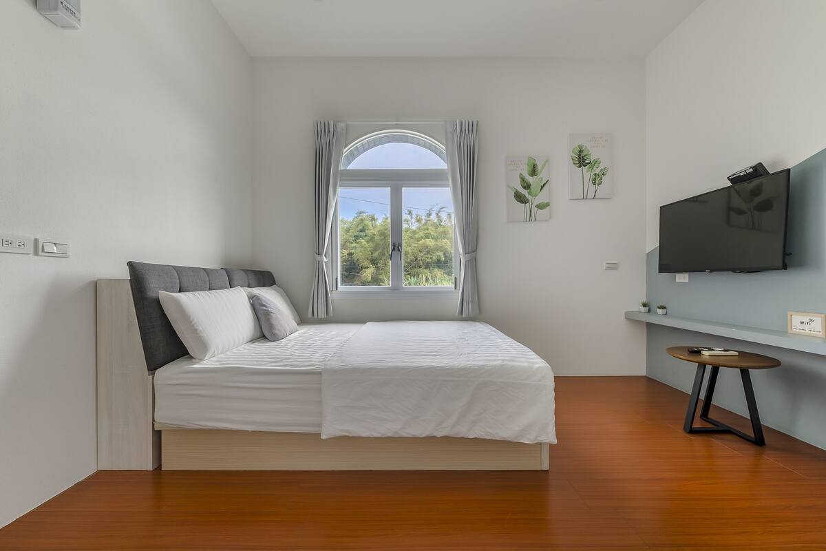 Quarto com cama, janela com cortinas, televisão na parede e uma pequena mesa.