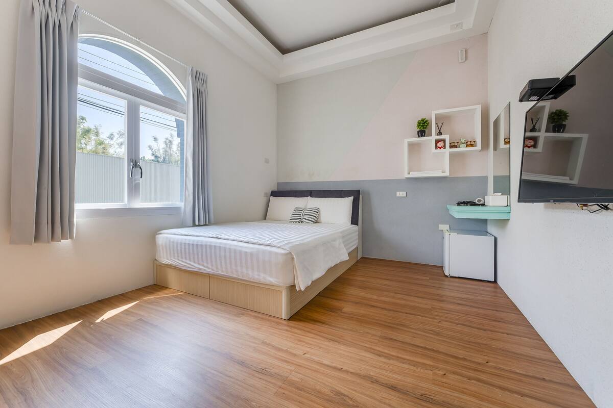 Quarto moderno com cama, mesa de cabeceira pequena, prateleira embutida e janela com cortinas.