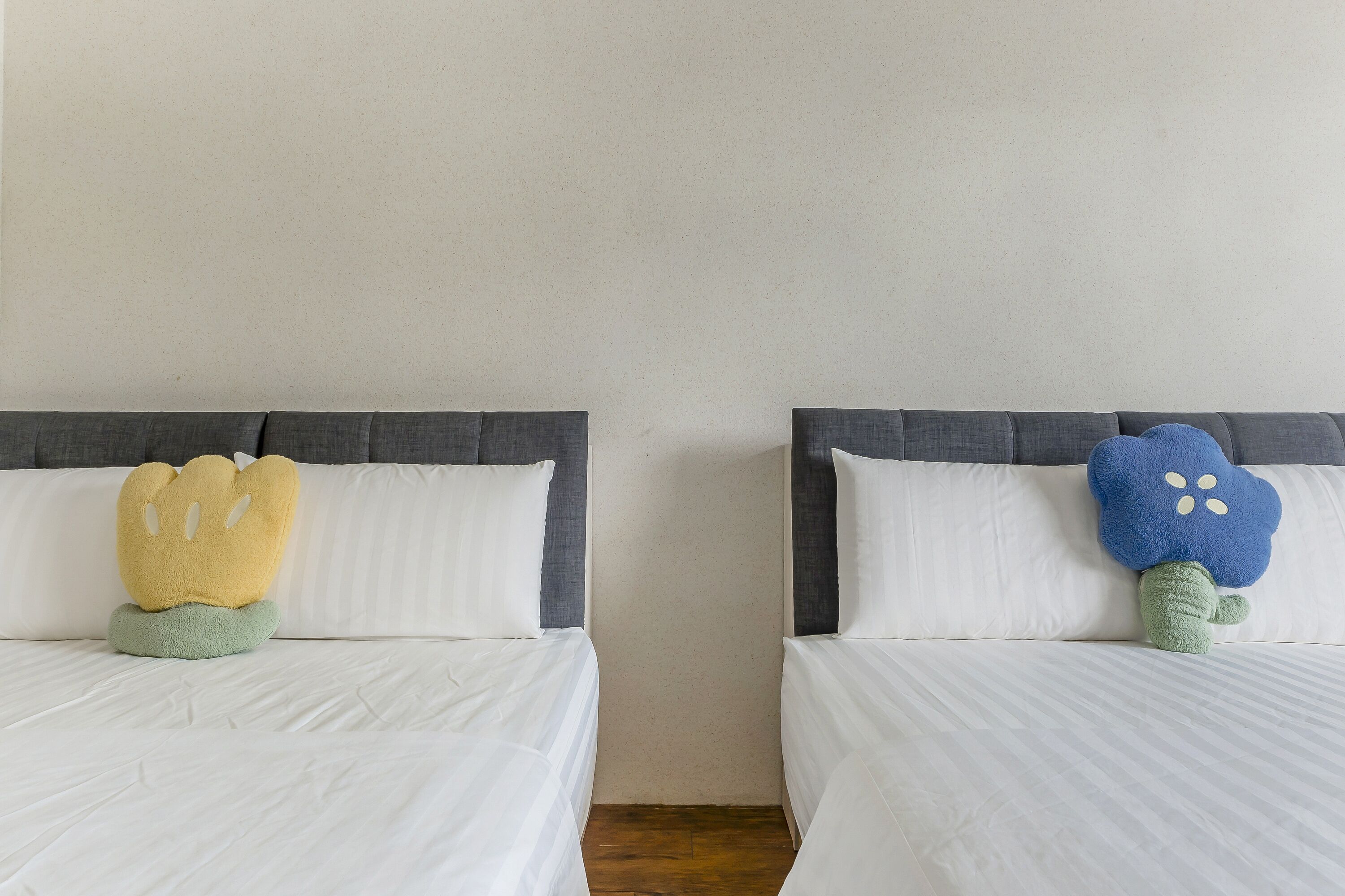 Dos camas con cabeceros grises y ropa de cama blanca, cada una con un cojín diferente.