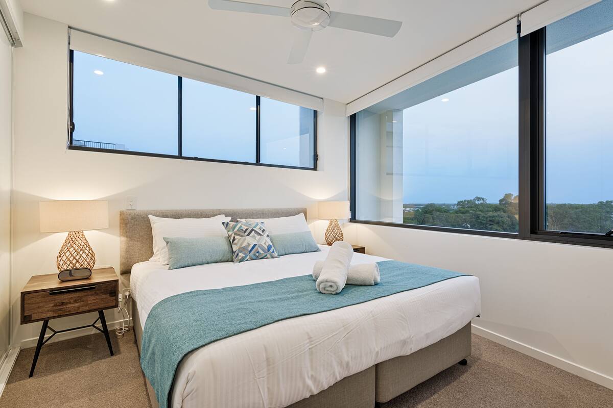 A modern bedroom with a large bed, two bedside tables, a ceiling fan, and large windows offering a view of the outdoors.