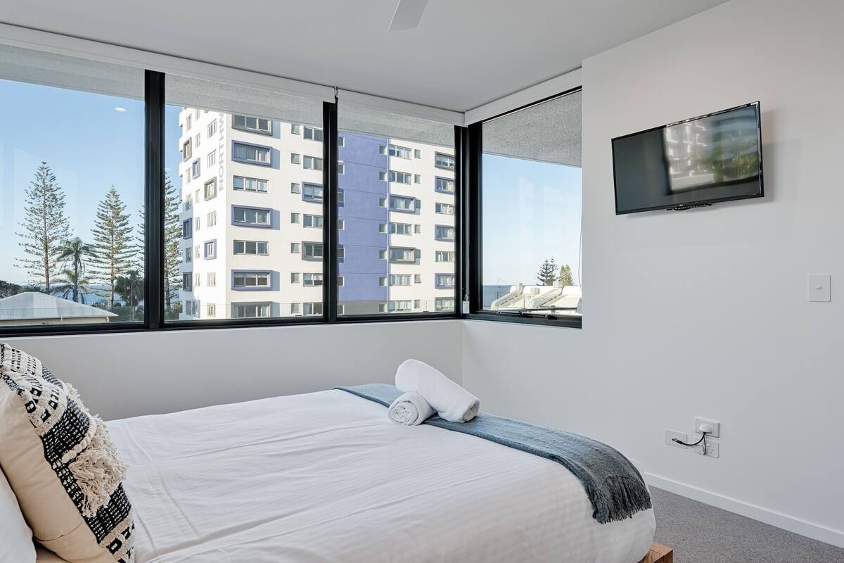A modern hotel room with a large bed, a TV mounted on the wall, and a view of a building through large windows.
