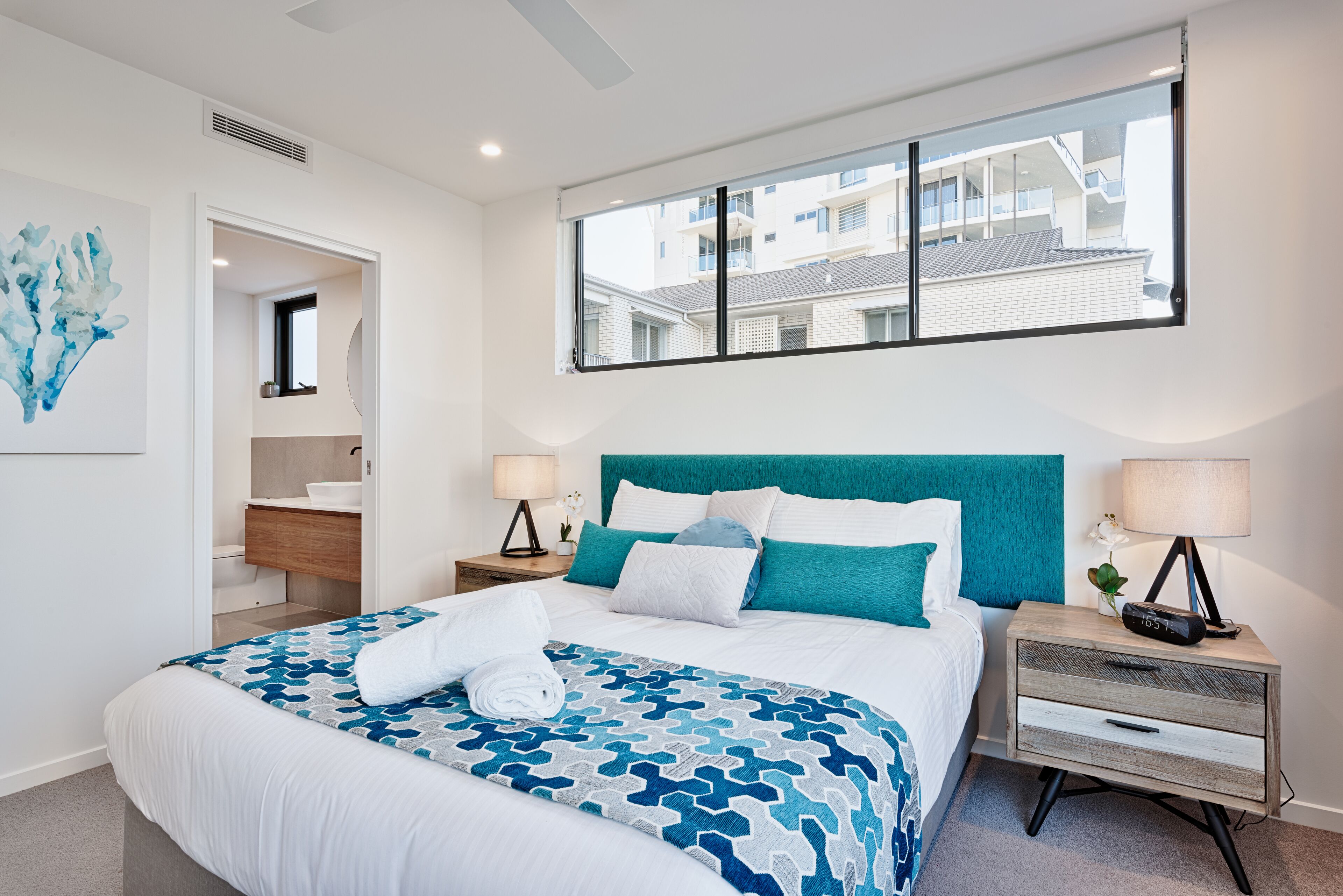 Una habitación de hotel moderna con una cama grande, cabecera color verde azulado, una mesita de noche con lámpara y vistas al edificio exterior.