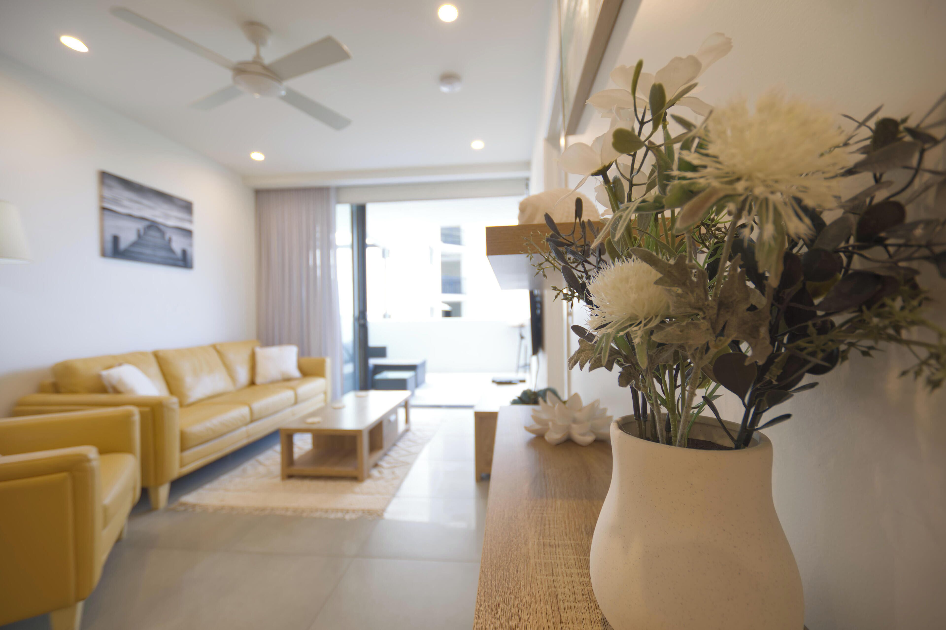 A modern living room with a yellow sofa, a wooden coffee table, and a white vase with flowers.