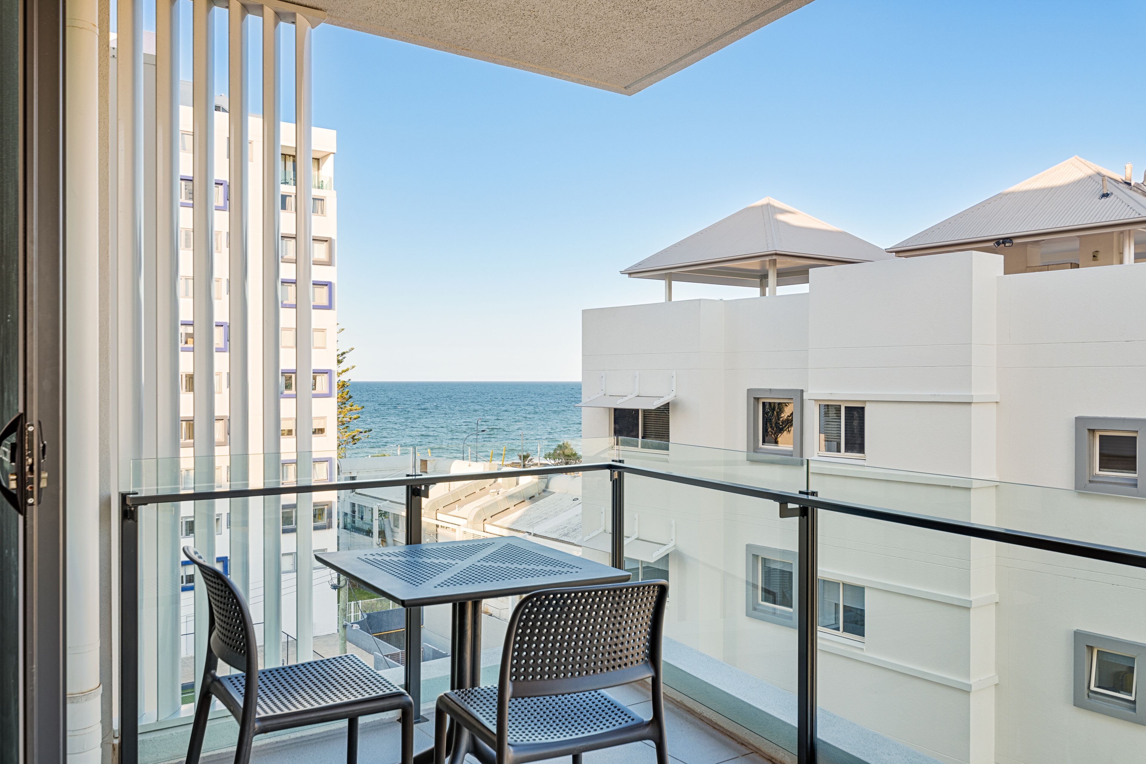 Un balcón con una mesa y dos sillas, con vista a una playa y edificios.