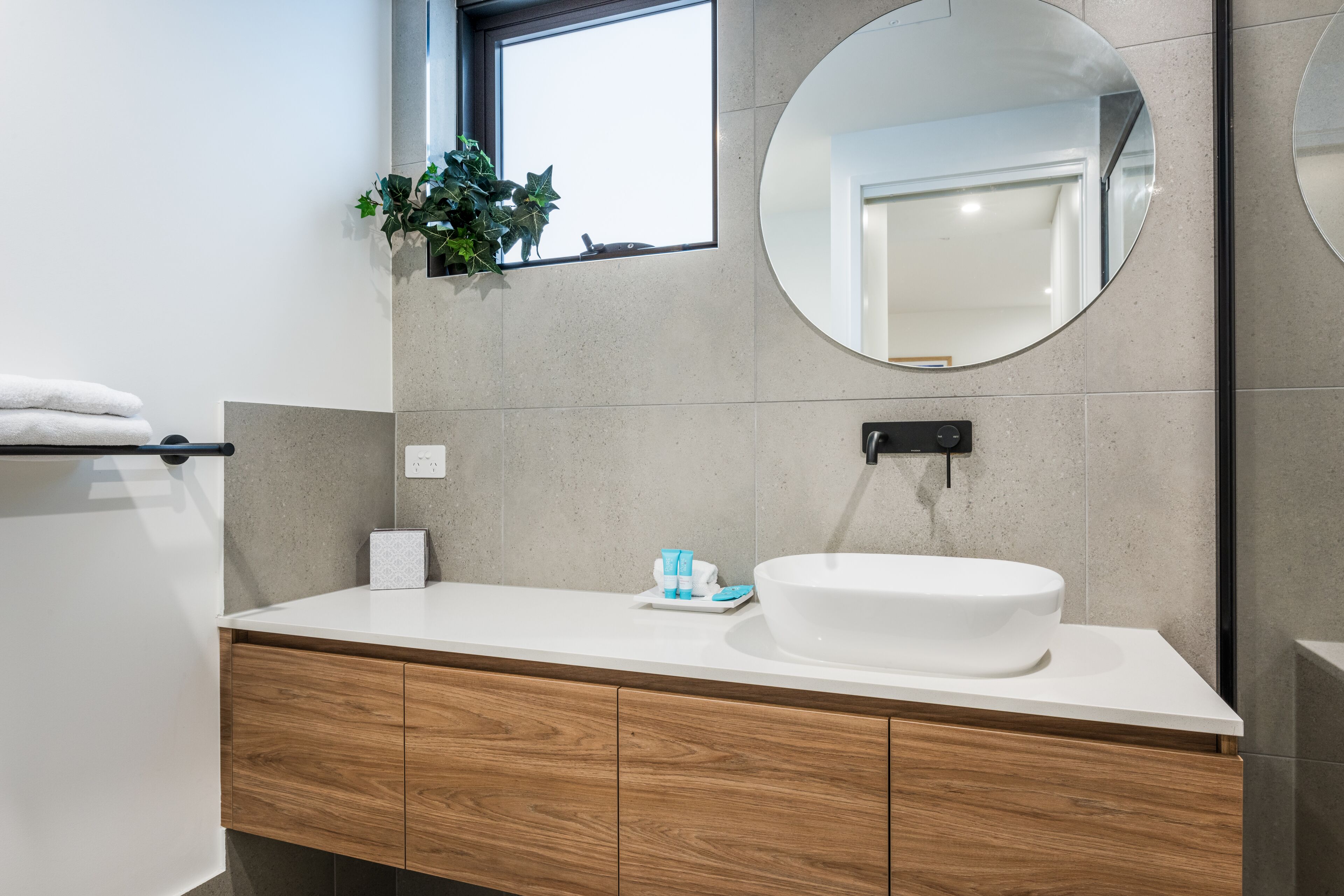 A modern bathroom with a white sink, wooden vanity, a round mirror, and a potted plant on the window sill.