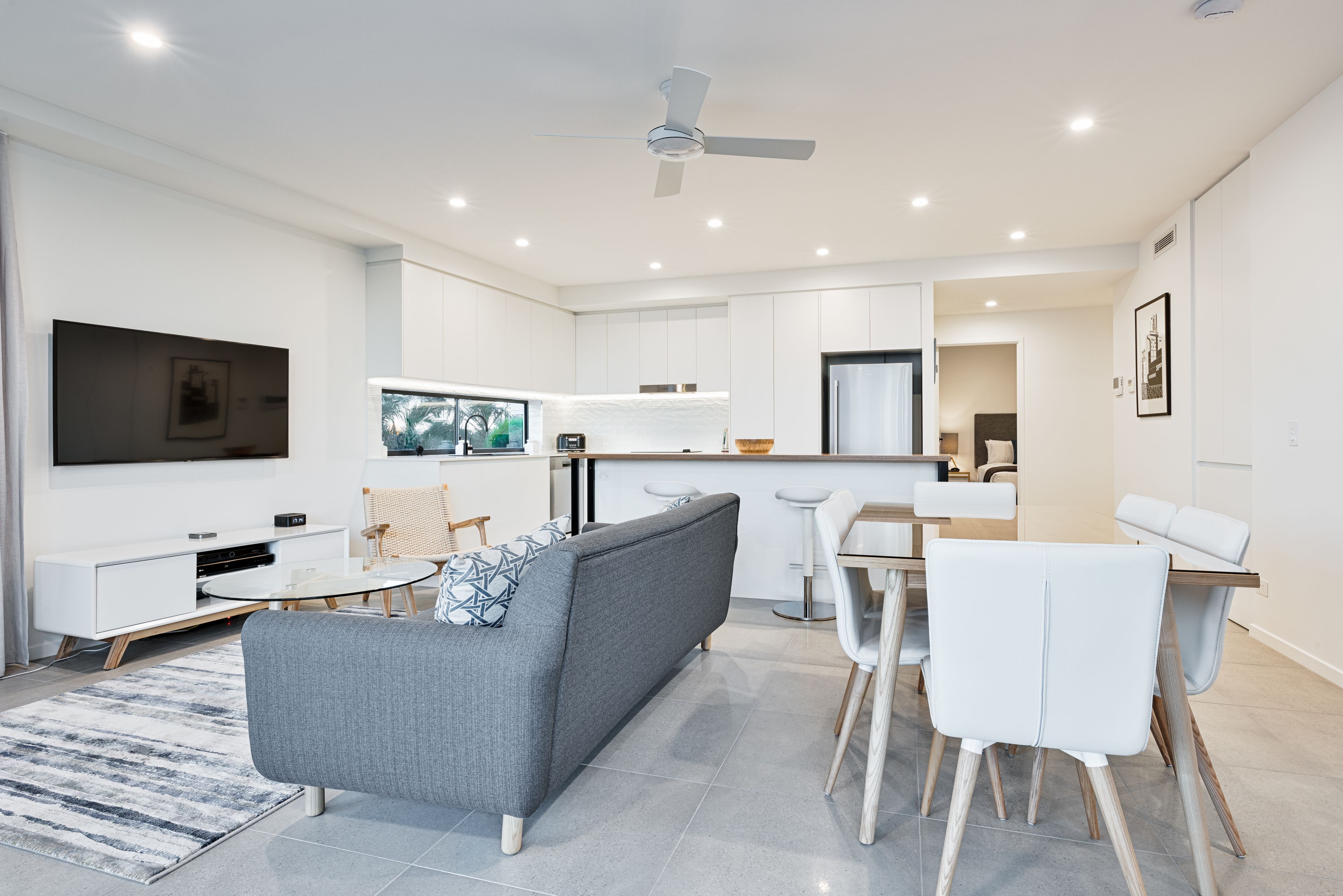 Uma sala de estar moderna com um sofá cinzento, uma mesa de jantar branca e cadeiras, uma televisão de tela plana e um ventilador de teto.