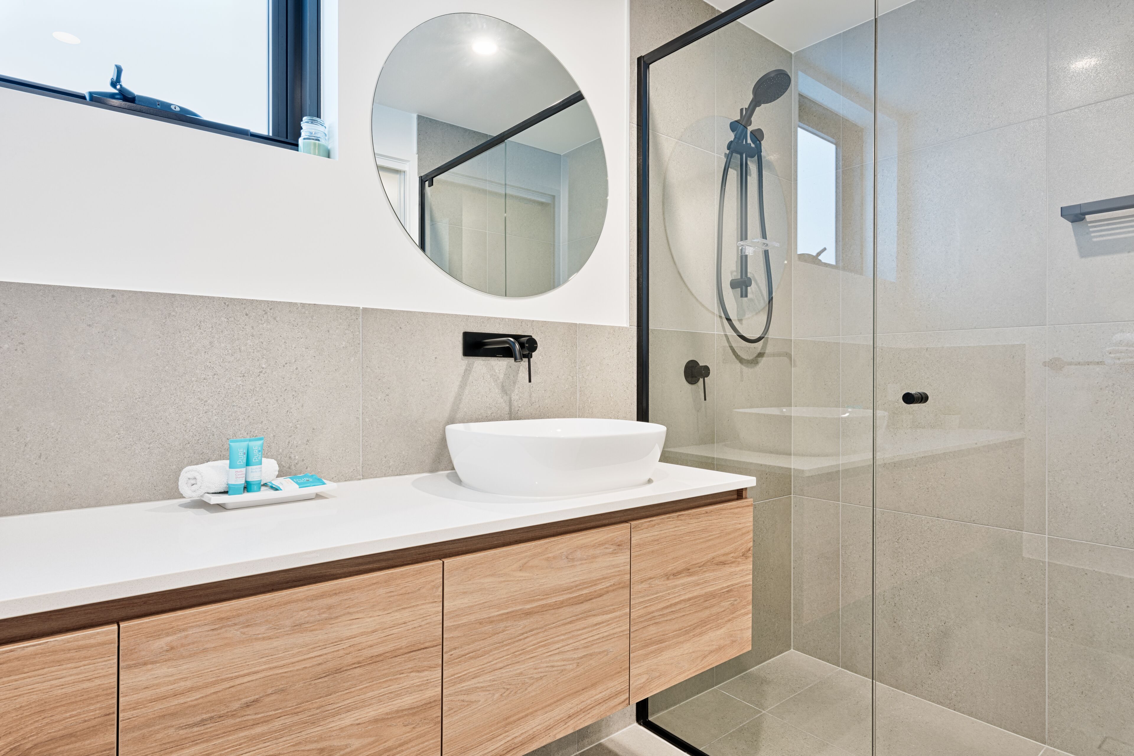 A modern bathroom with a white sink, wooden cabinets, a round mirror, and a glass-enclosed shower area.