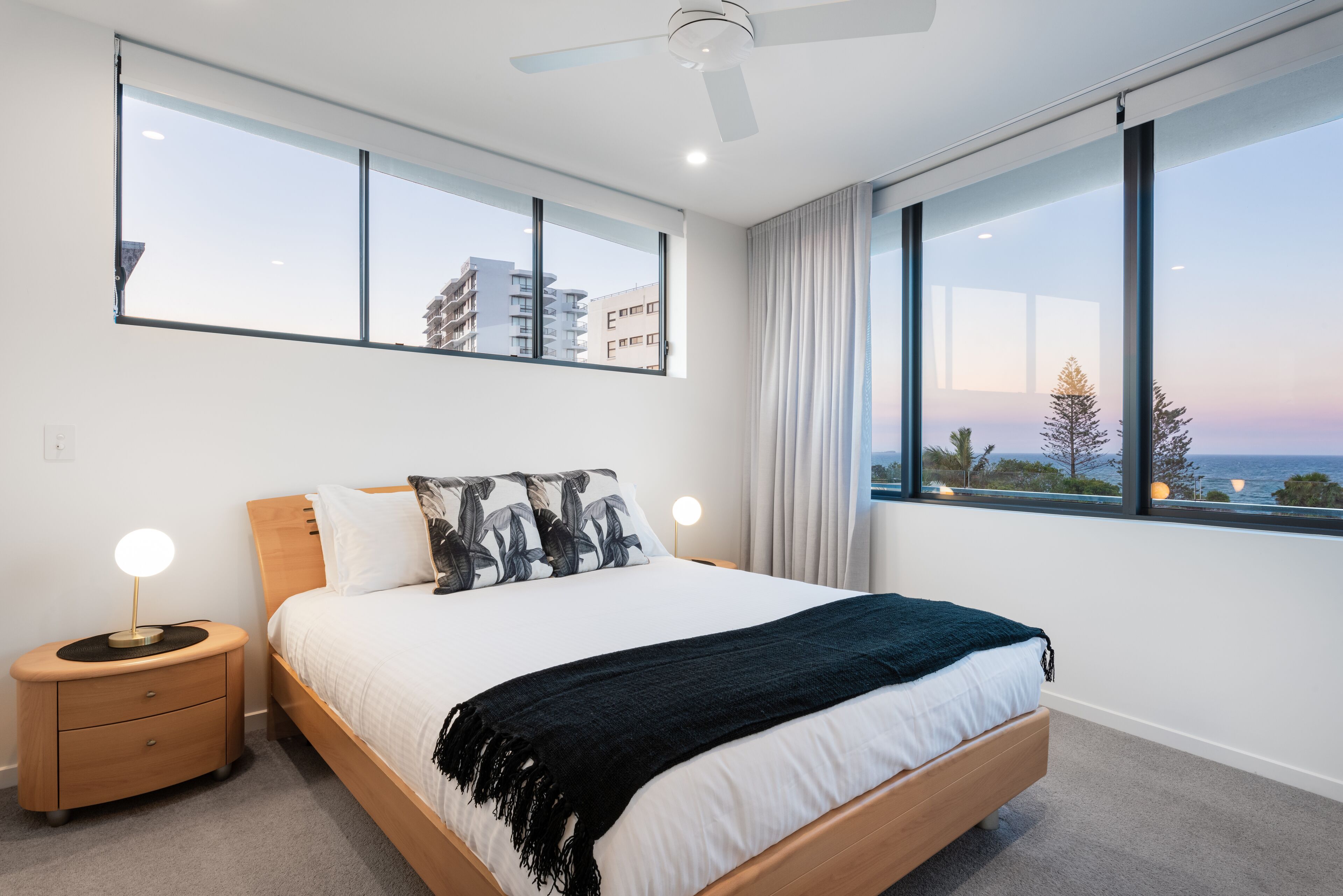 Een moderne slaapkamer met een groot bed, twee nachtkastjes, een plafondventilator en grote ramen met uitzicht op gebouwen en de zee.