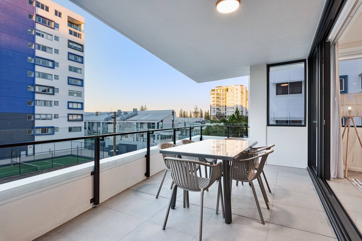 A balcony with a dining table and chairs, overlooking a tennis court and surrounding buildings.