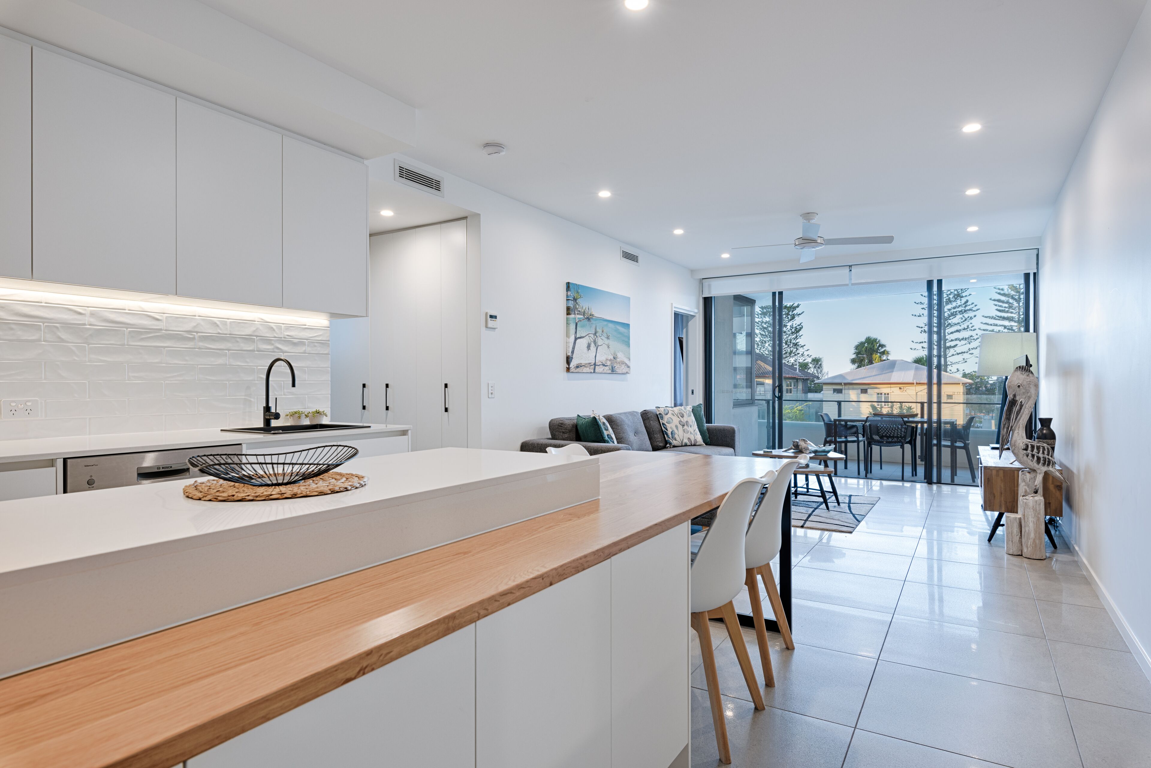 Una cocina moderna con una isla central, armarios blancos y un comedor con puerta de vidrio que da a un balcón.