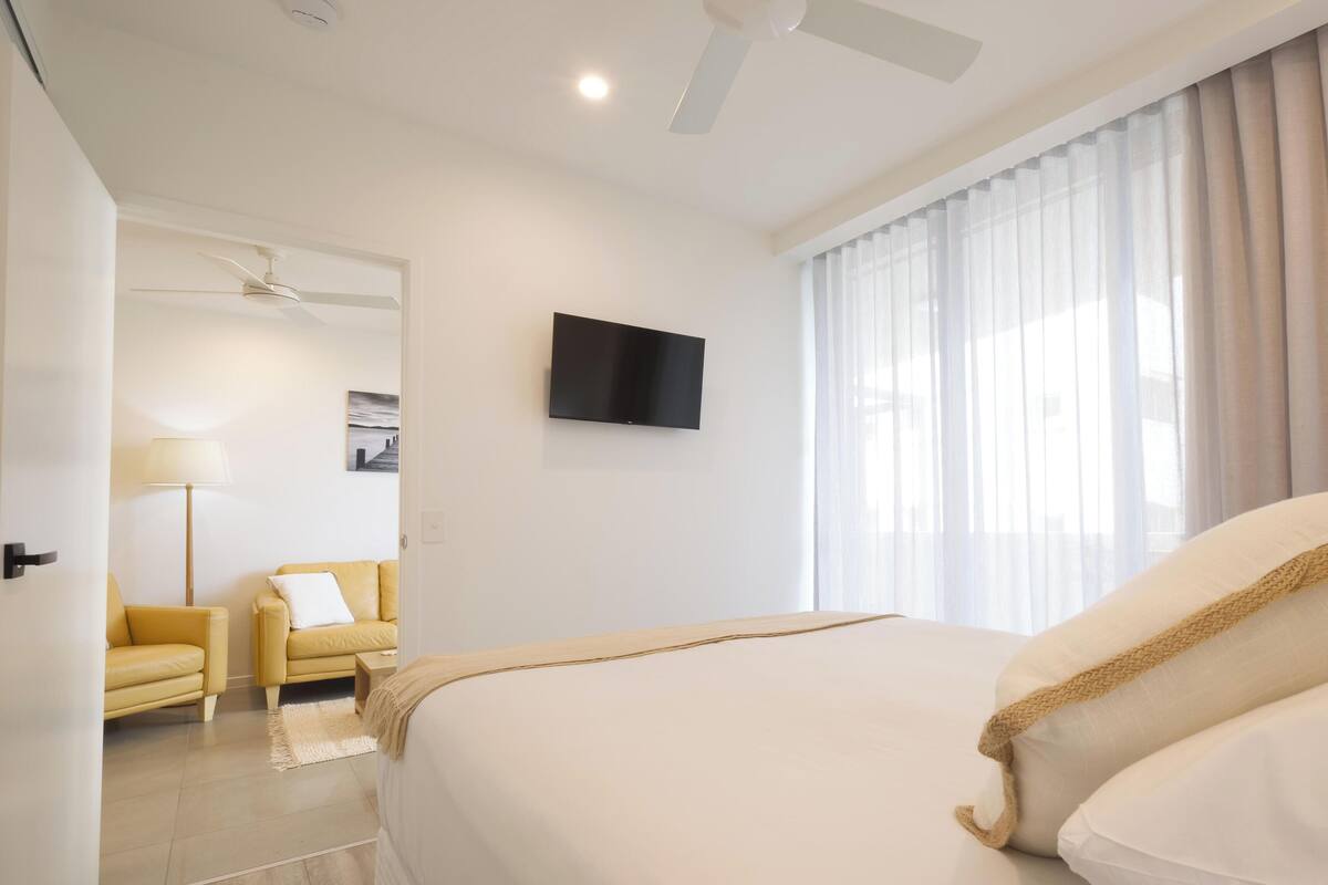 A hotel room with a bed, a TV mounted on the wall, a ceiling fan, and a window with curtains.
