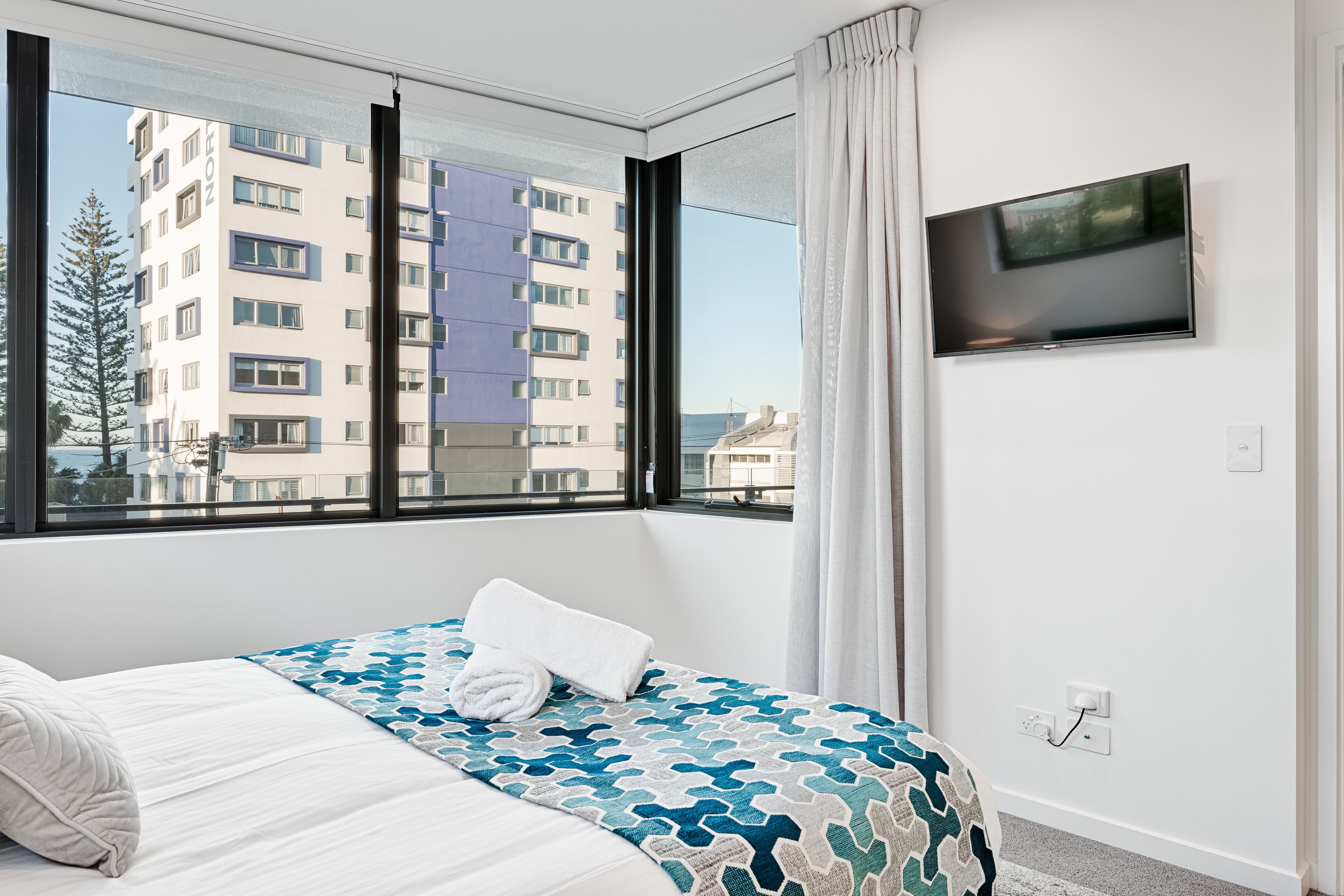 A hotel room with a large window, a bed with a patterned bedspread, a flat-screen TV mounted on the wall, and a view of the cityscape.