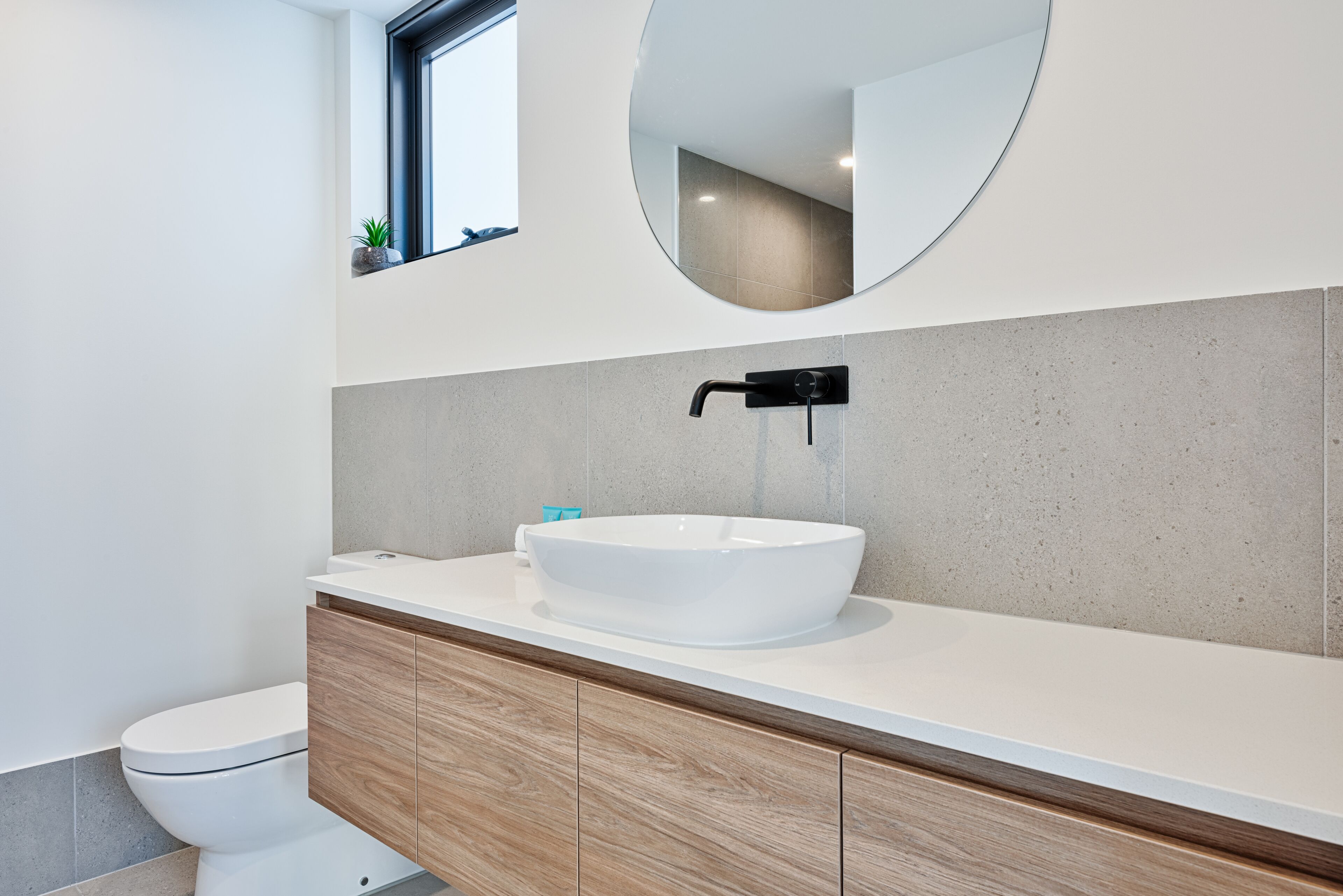 A modern bathroom with a white sink, wooden vanity, a round mirror, and a toilet.