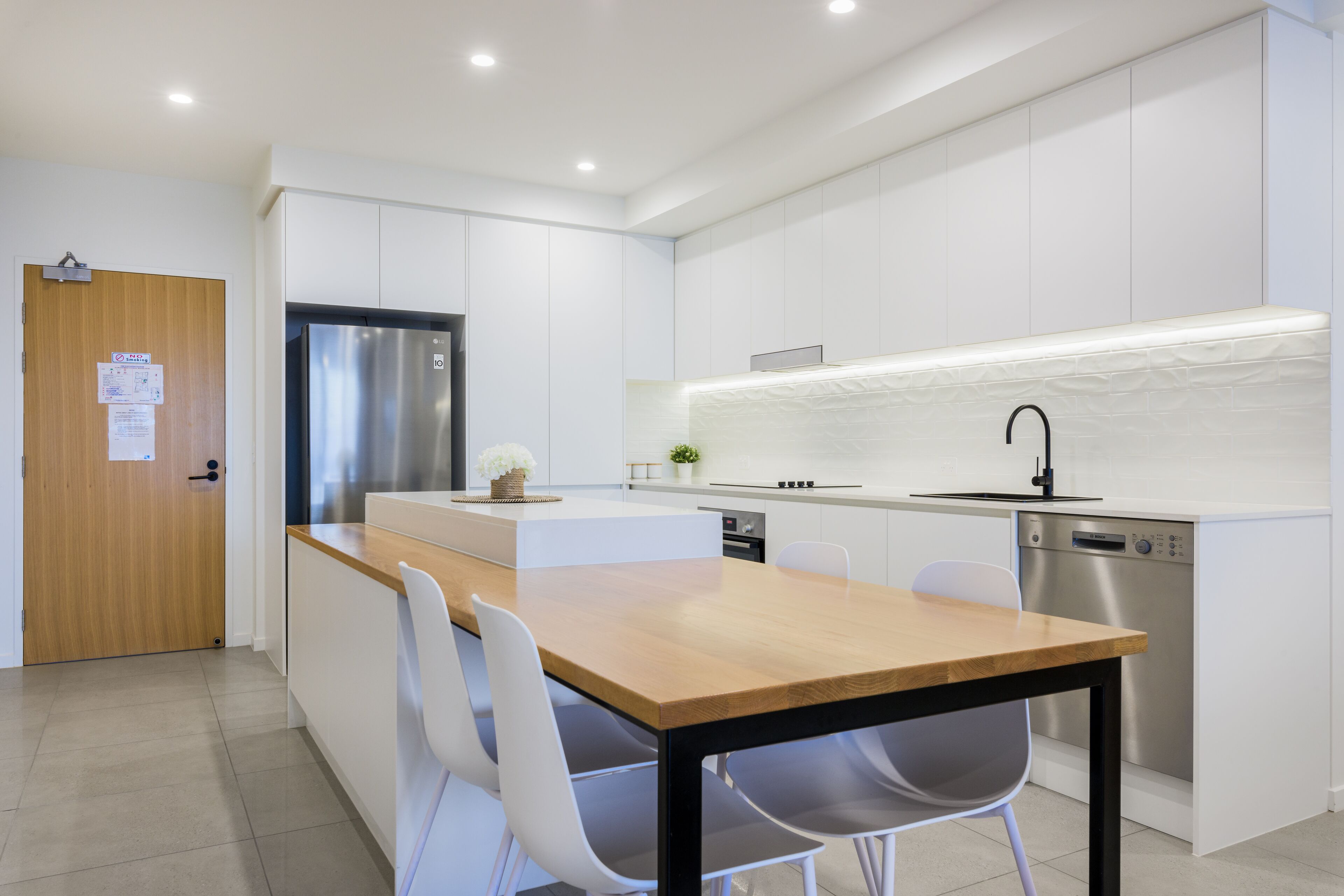 A modern kitchen with white cabinetry, a stainless steel refrigerator, a wooden dining table with chairs, and built-in appliances.