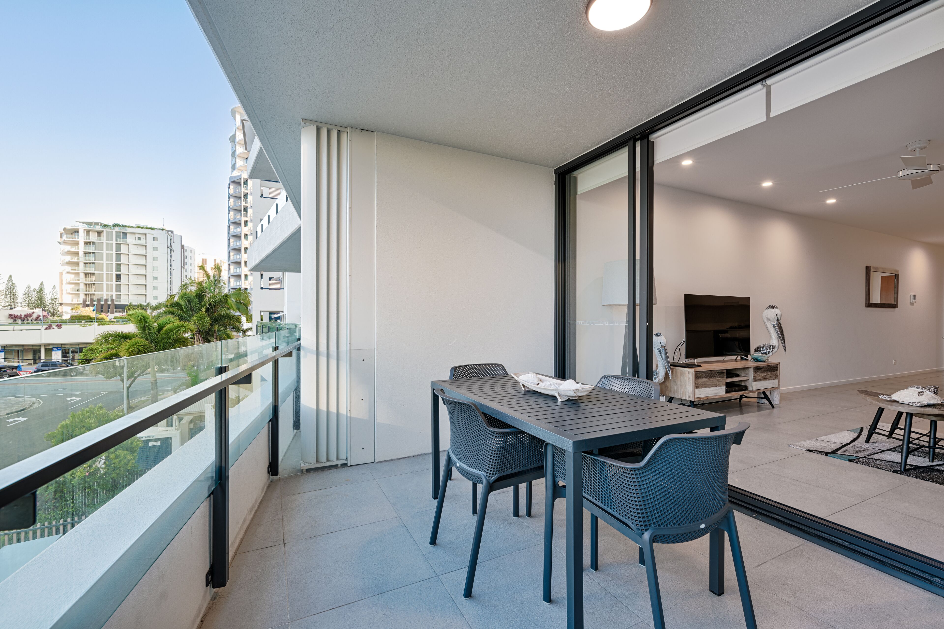Ein Balkon mit Tisch und Stühlen, ein Fernseher im angrenzenden Raum und Blick auf eine Stadtlandschaft.