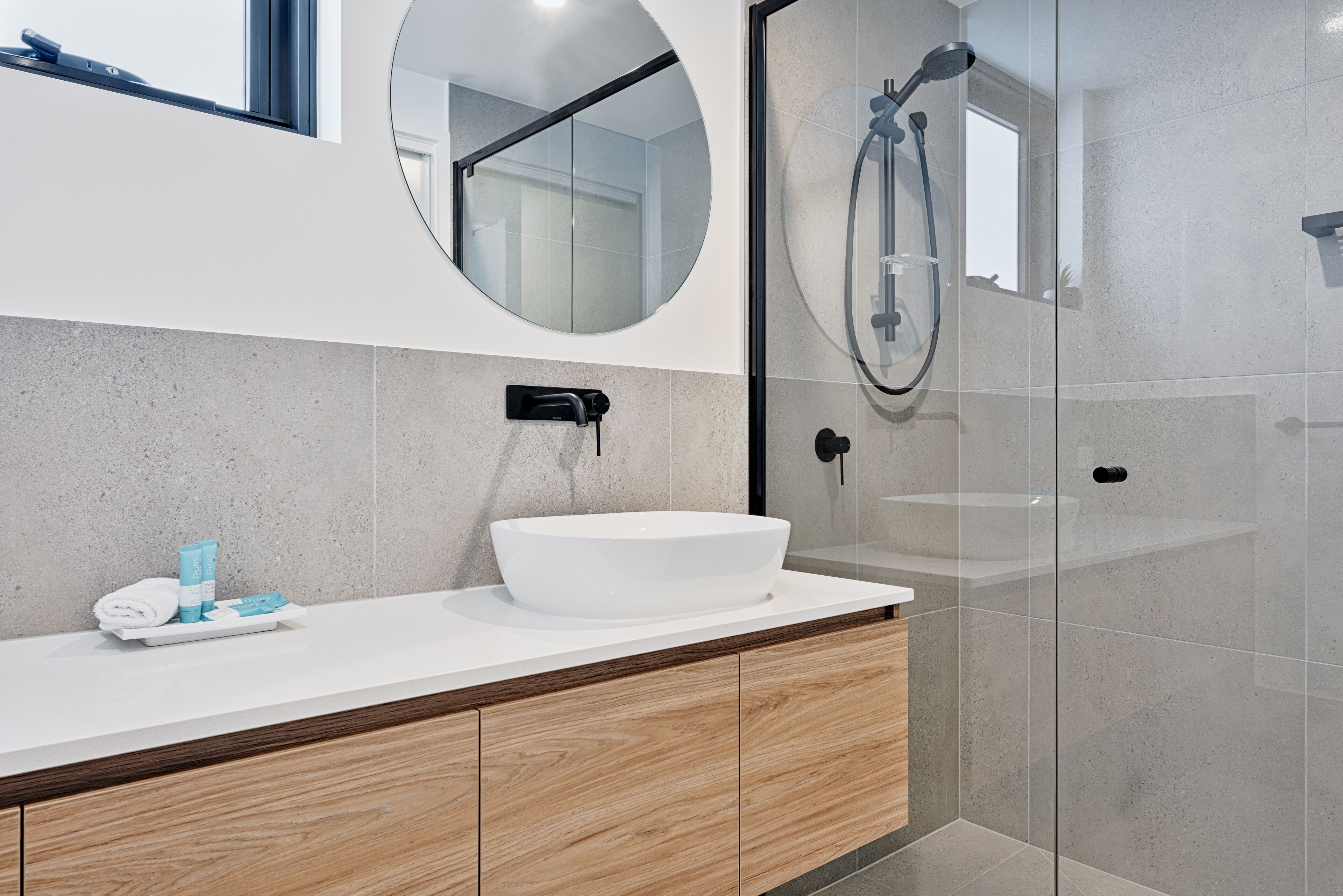 A modern bathroom with a white sink, wooden vanity, a round mirror, and a glass-enclosed shower area.