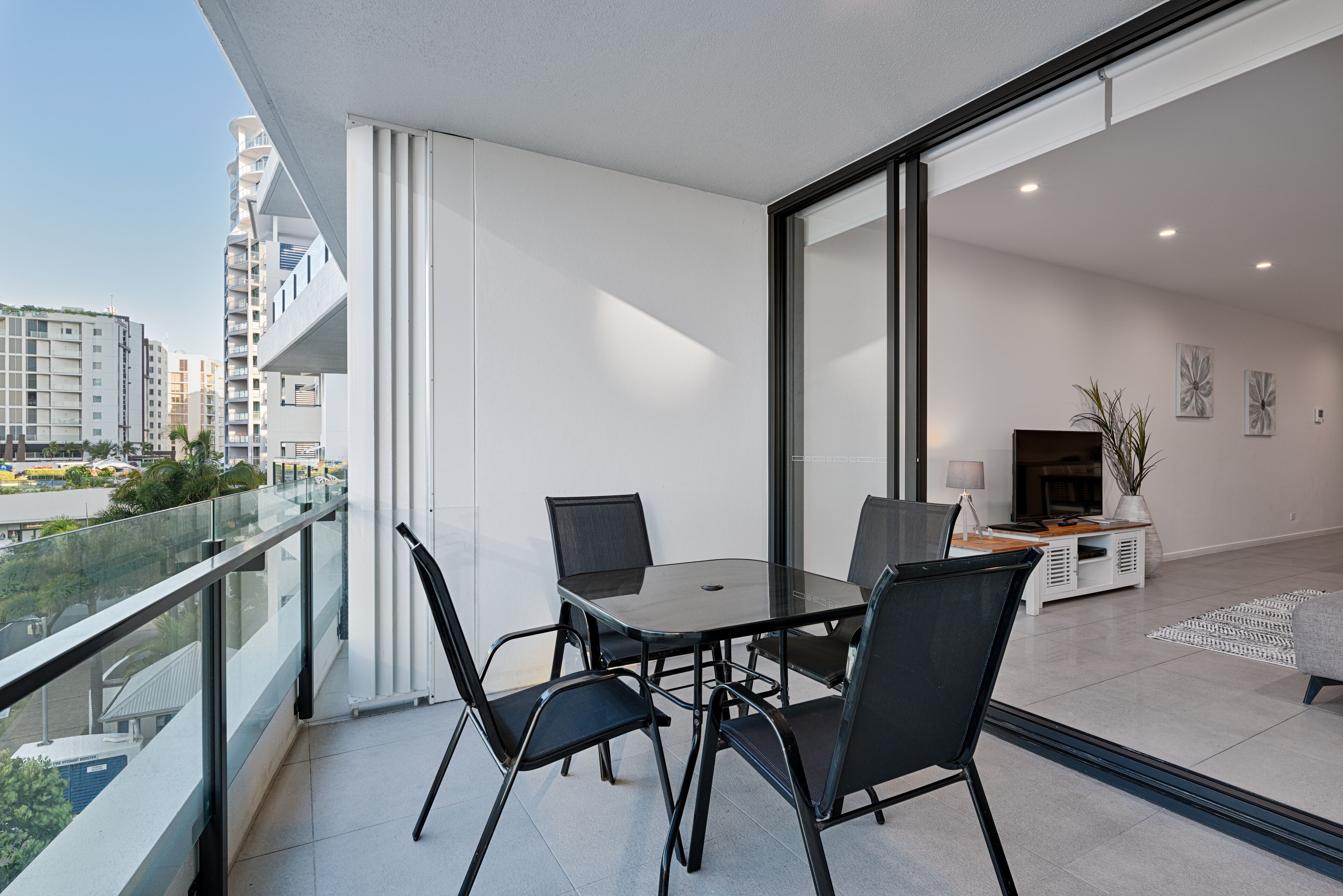 Un balcón moderno con barandilla de vidrio, una mesa de comedor con sillas y vistas a un paisaje urbano.