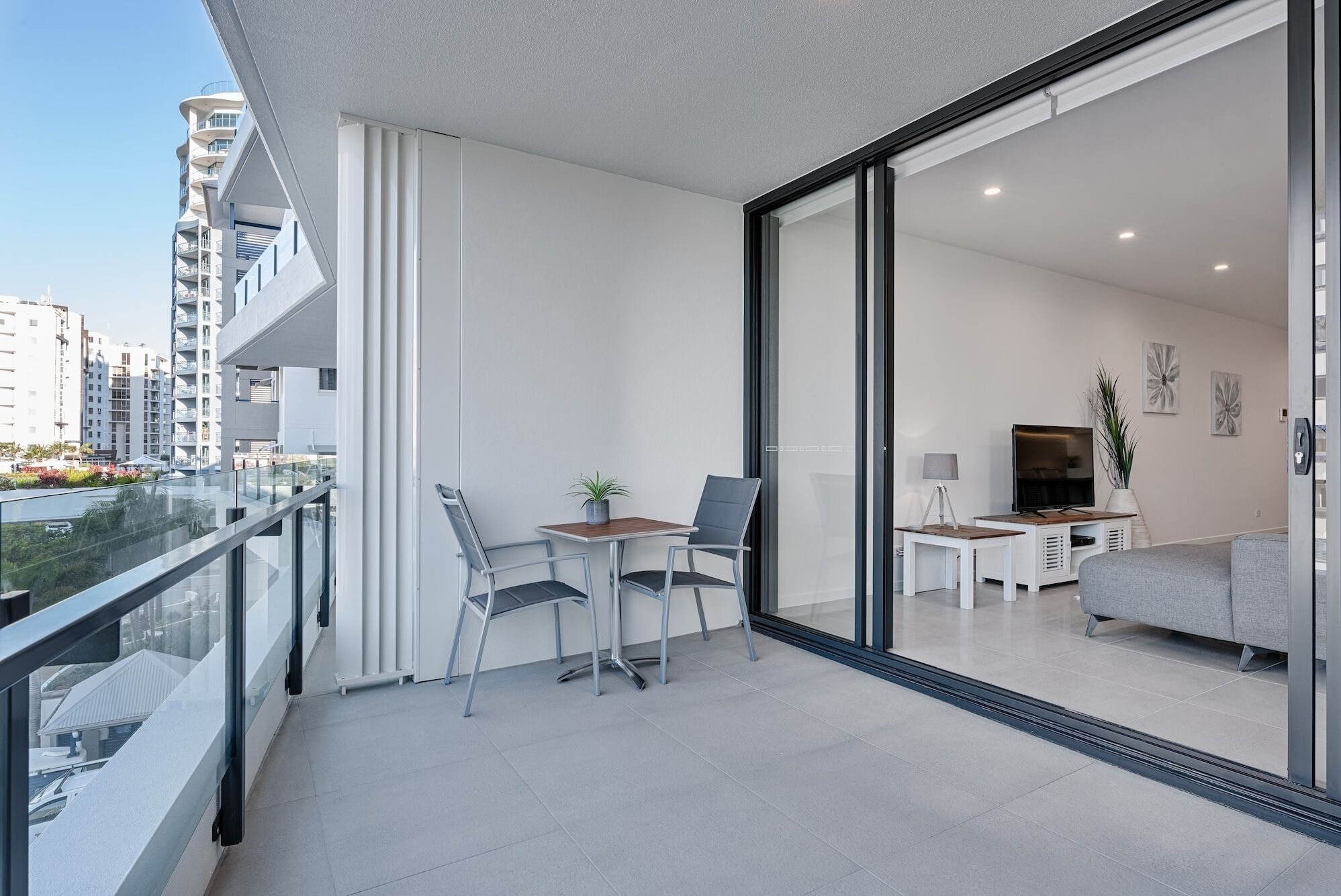A modern apartment with a balcony, a dining area with two chairs and a table, a living room with a sofa and a TV, and a large sliding glass door.