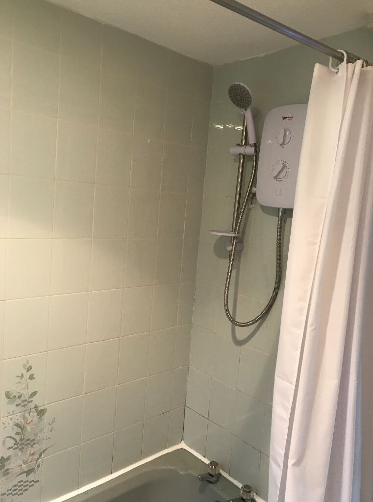 Un petit espace douche fermé, doté d’un pommeau de douche à main et d’un panneau de commande fixé au mur.