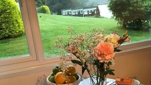 A table set with a cake, fruit bowl, and a vase of flowers, with a view of a house and greenery outside. - Welcome to Poldark Country - A Cornish Idyll! (Camelford)
