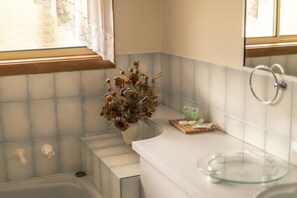 Un baño con un lavamanos blanco, un cuenco de vidrio, un florero con flores secas y una ventana con cortinas de encaje.