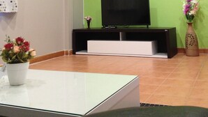 Un soggiorno con una TV a schermo piatto su un supporto nero, un tavolino da caffè bianco, una pianta in vaso e un vaso con fiori.