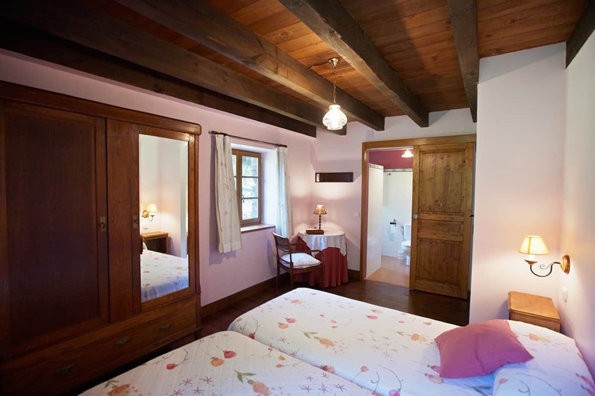 A bedroom with two beds, wooden wardrobe, a small table with a lamp, and a door leading to another room.