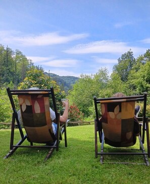 Duas cadeiras de exterior com encosto estampado, dispostas sobre um relvado com vista para uma encosta arborizada.
