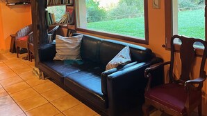 A cozy living room with a black leather sofa, a wooden chair, a bookshelf, and a large window offering a view of greenery. - Casa Rural Goikola (Deba)