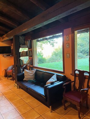 Uma sala de estar acolhedora com um sofá de couro preto, uma cadeira de madeira, uma estante de livros e uma janela grande com vista para o verde.