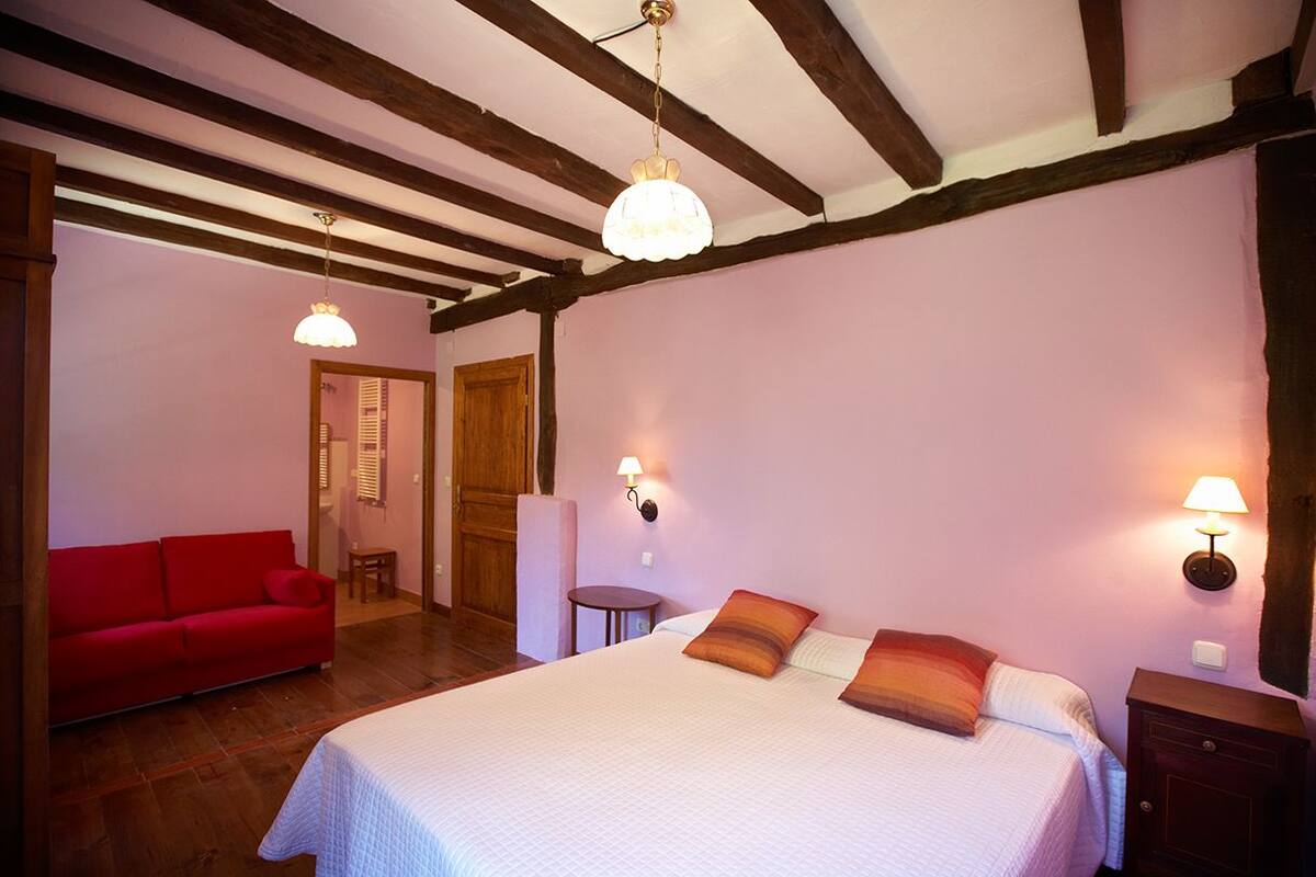 A bedroom with a large bed, a red sofa, wooden beams on the ceiling, and two wall-mounted lamps.