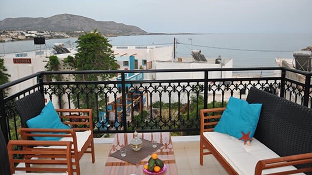 A balcony with two wooden chairs and a table, overlooking a coastal view with buildings and a mountain in the distance. - Porfyra's Island (Ierapetra)