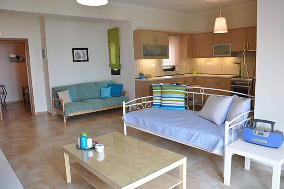 A modern living room with a sofa, a bed, a glass coffee table, and a kitchen area with cabinets and appliances.