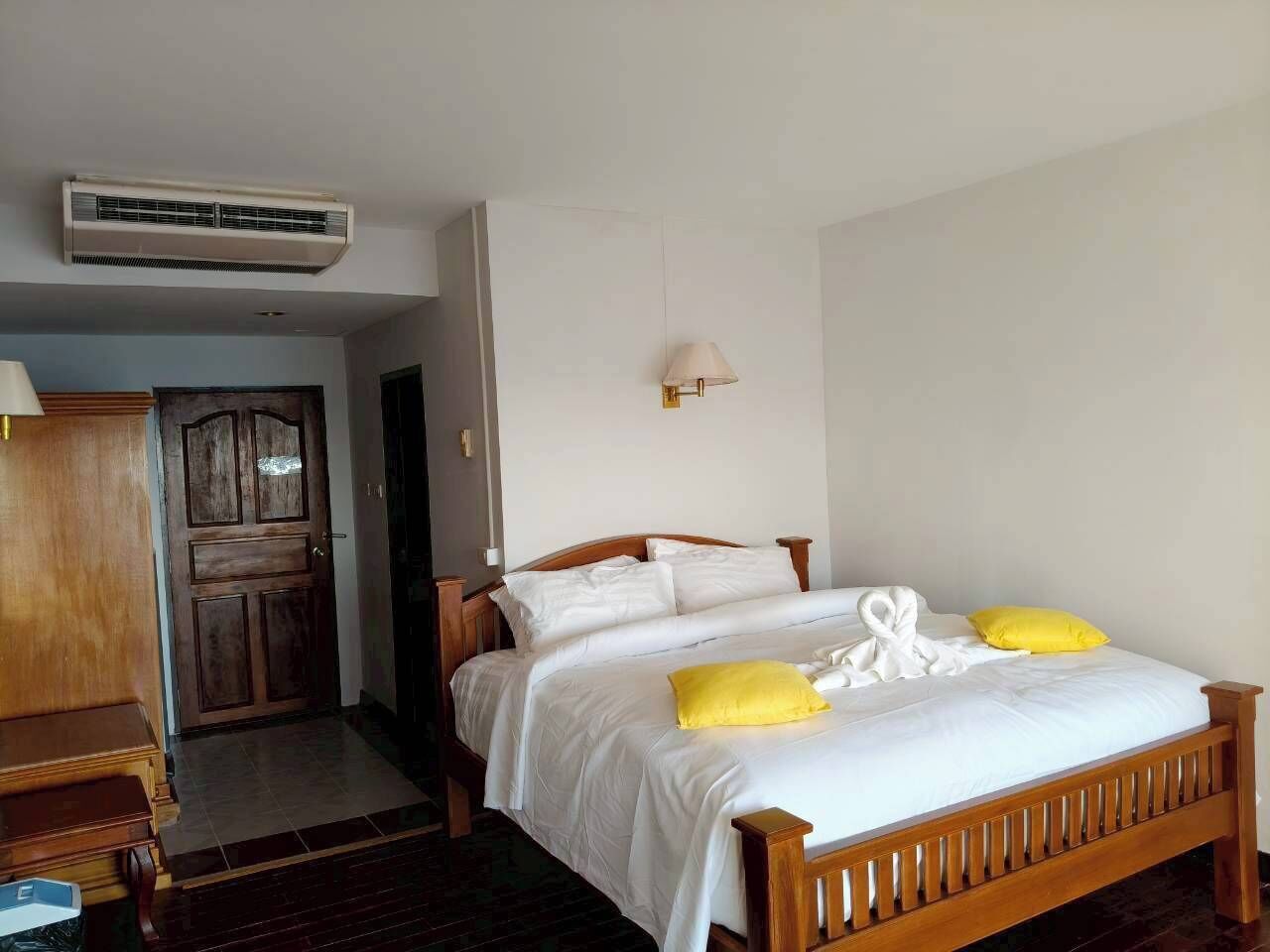 A bedroom with a wooden bed, white bedding, a yellow pillow, a white towel, and a wall-mounted lamp.