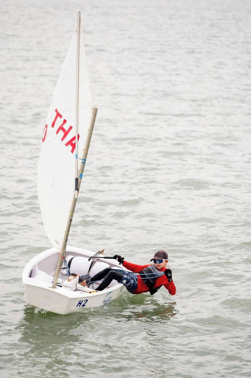 A person on a sailboat with a white sail marked "THA" and the number "H2".