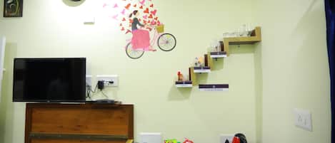 A hotel reception area with a flat-screen TV, a clock, a wall decal of a woman on a bicycle, and shelves labeled 'Cafeteria' and 'Shop'.