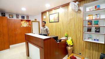 A reception area with a wooden reception desk, a clock, a fan, a bookshelf with books, and a flower vase.