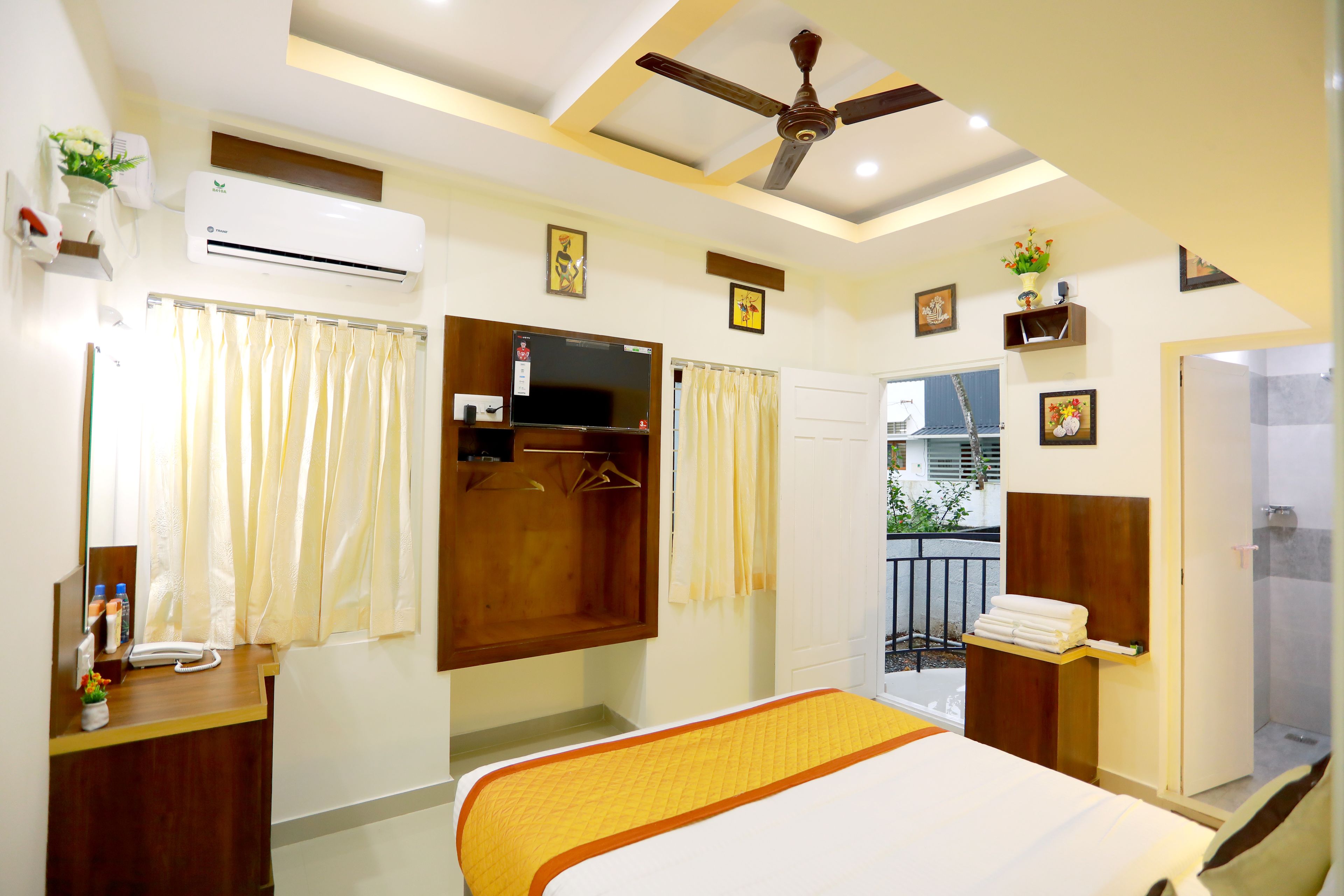 A hotel room with a bed, a desk, an air conditioner, a ceiling fan, a shower area, and a balcony with a railing.