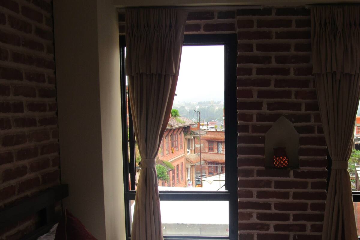 Quarto de hotel com cama, cortinas, parede de tijolos e vista para edifícios.
