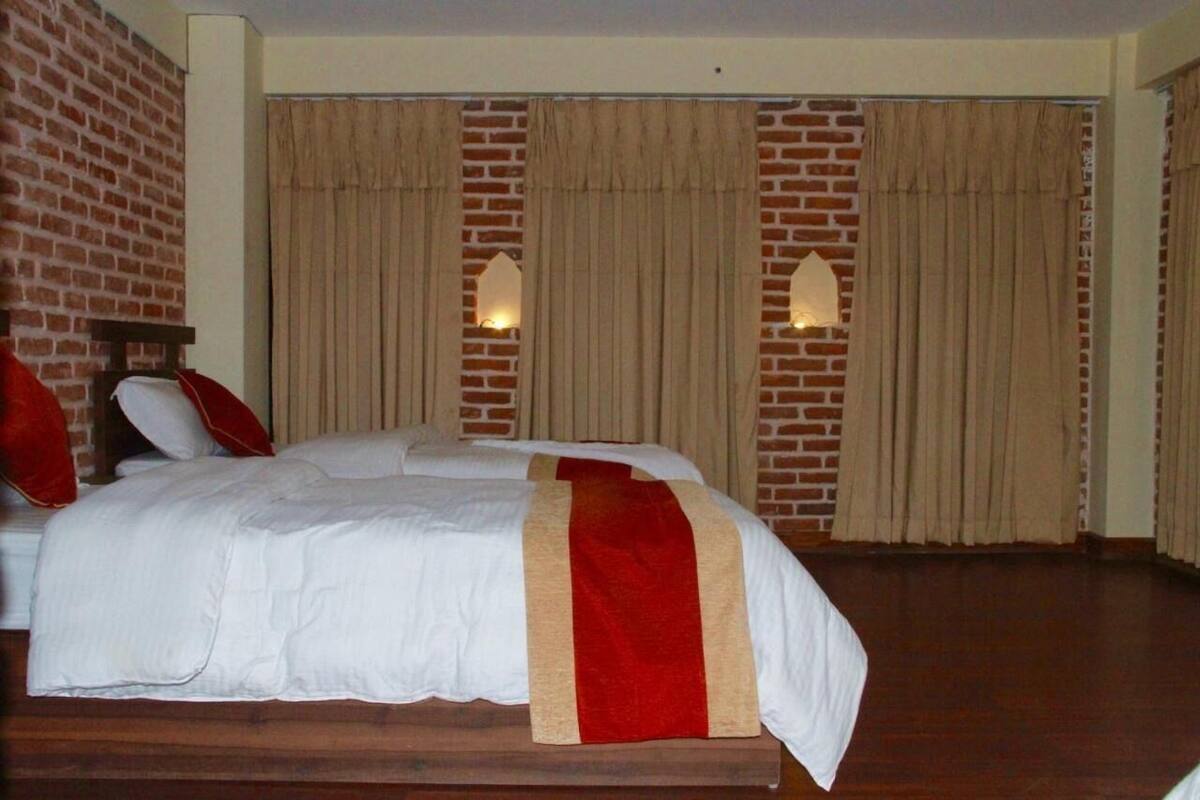 Quarto de hotel com parede de tijolos, uma cama grande com roupa de cama branca e um toque de vermelho, e duas luzes fixadas na parede.